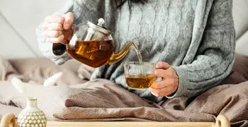 Woman in sweater pours tea into a mug while sitting in bed stock photo